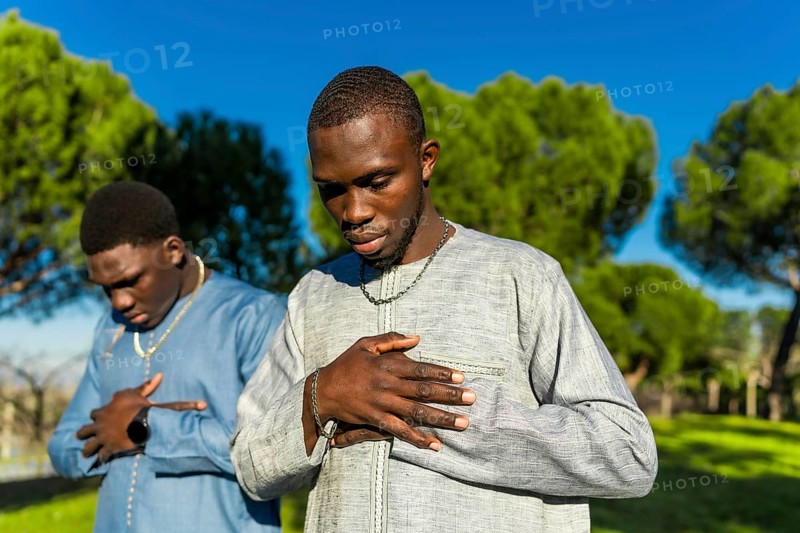 Two young senegalese men wearing traditional clothing are praying outdoors in a park, expressing their faith and devotion
