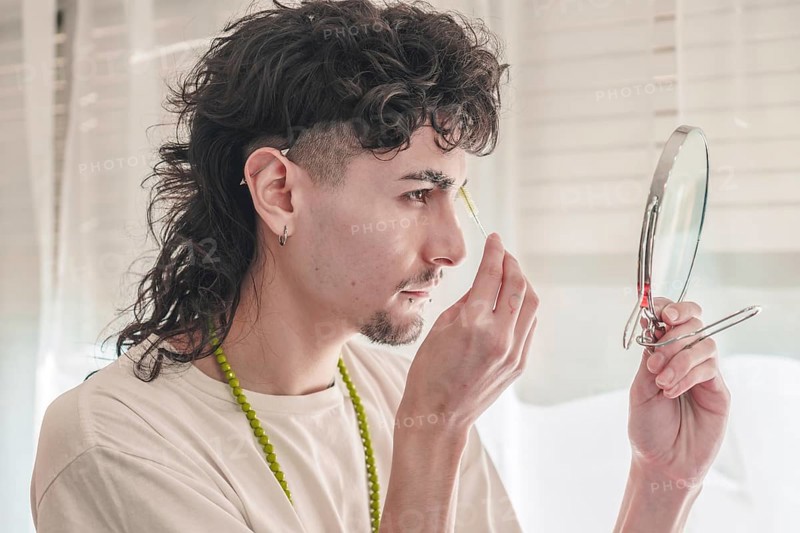 Concentrated young man with curly hair plucking his eyebrows with tweezers while looking in a small mirror, taking care of his appearance in a bright room