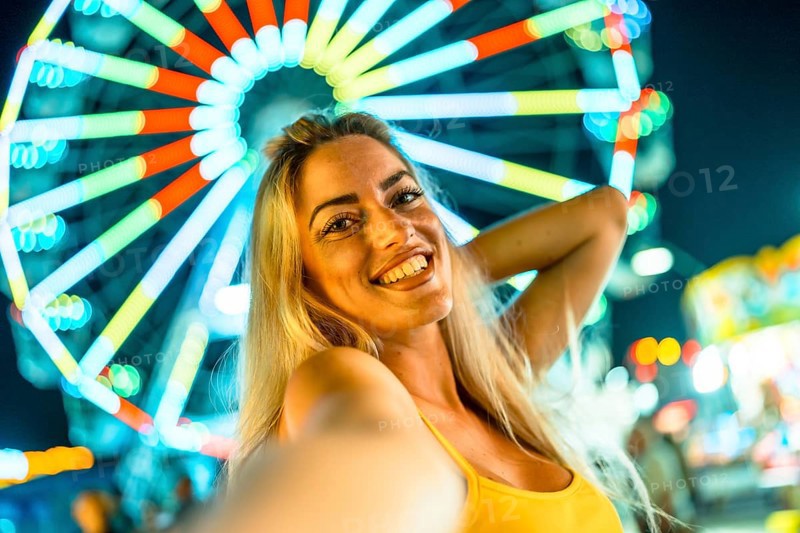 Personal point of view of a beauty happy woman taking selfie in an amusement park at night with a big wheel on the background illuminated by neon lights