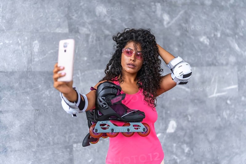 Latin woman in sportswear and protective gear taking selfie holding inline skates outdoors