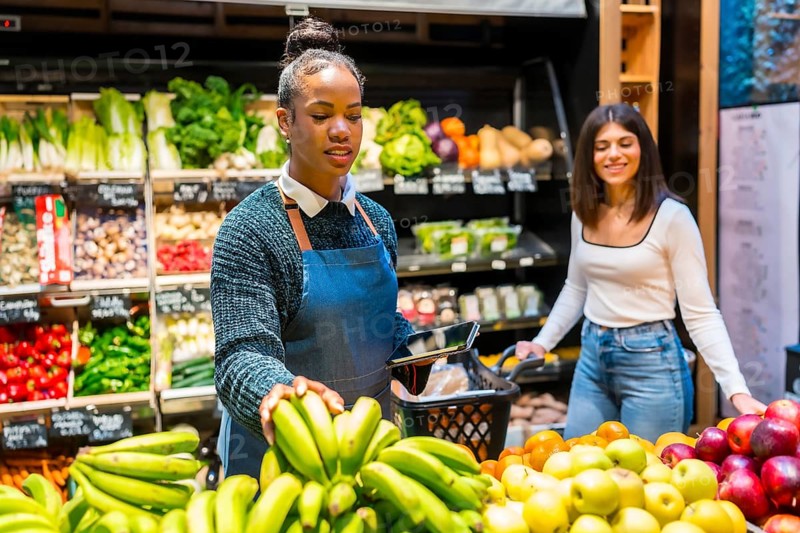 Greengrocer arranging fresh bananas while using digital tablet and helping customer in organic supermarket with colorful fruit and vegetables