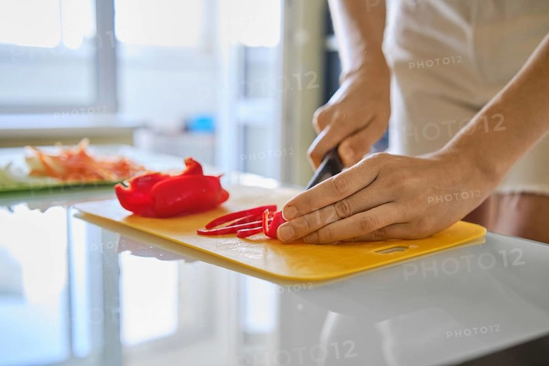 A person is slicing fresh red bell peppers on a yellow cutting board in a well-lit kitchen