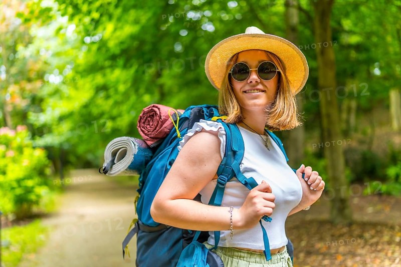 Cheerful caucasian young woman wearing sunglasses and sun hat carrying backpack walking along a path enjoying nature in the forest
