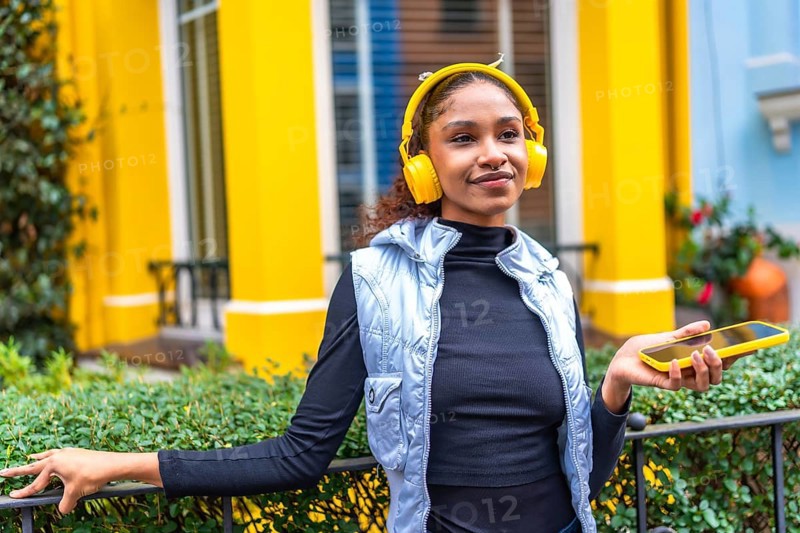 Afro woman listening to music relaxes in the city leaning on garden rail of a colorful yellow house