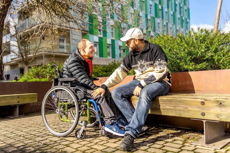 Two friends talking and smiling on a bench in a city park, one of them is using a wheelchair