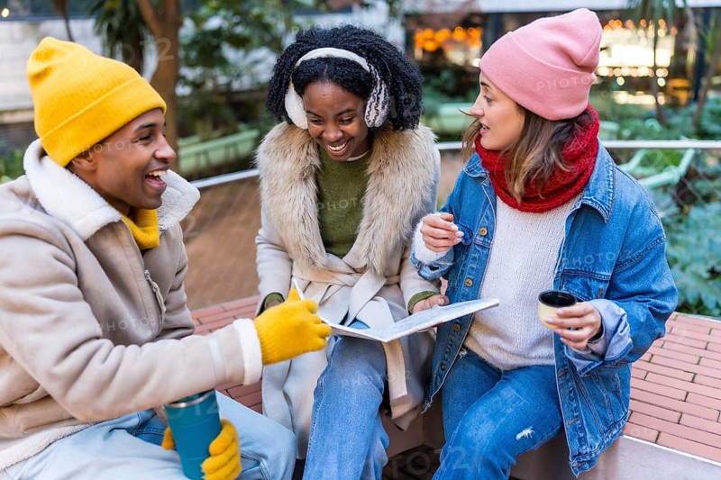Three cheerful young students are sitting on a bench, drinking coffee and studying together on a cold winter day in the city