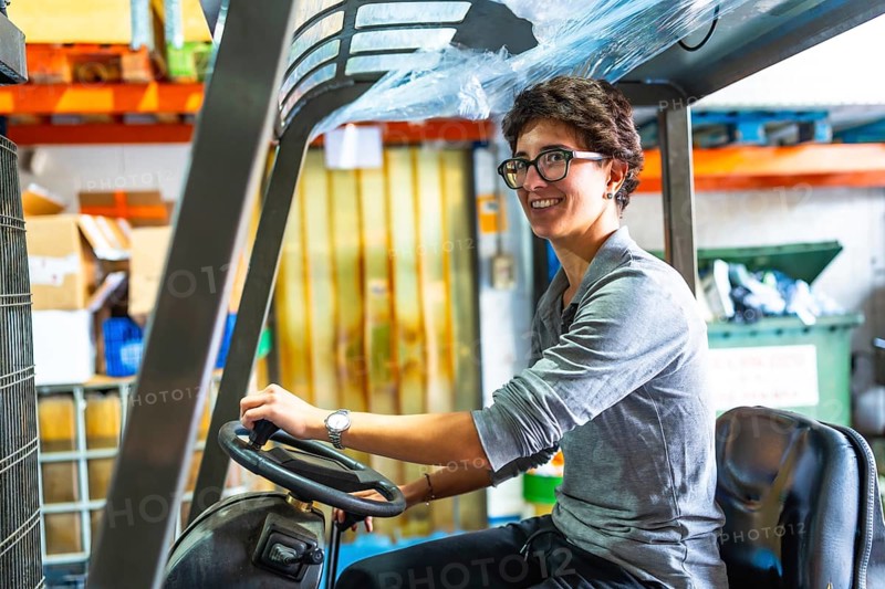 Side view of a happy caucasian woman with short hair and eyeglasses driving forklift in a metallurgic factory