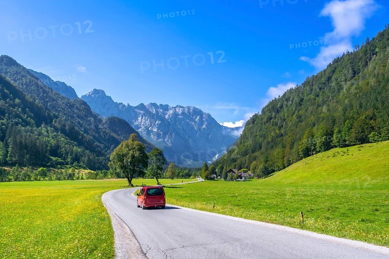 Red van driving along a winding road in the picturesque logarska dolina valley, surrounded by lush meadows, towering mountains, and a clear blue sky