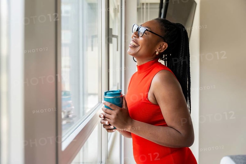 Happy businesswoman holding reusable coffee cup and looking out window during business trip, enjoying city view