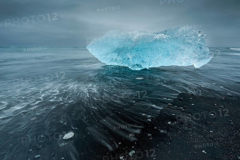 A shimmering blue iceberg is washed up by waves on a dark beach, Diamond Beach, Jökulsárlón glacier lagoon or Jökulsarlon, South Iceland, Iceland, Europe