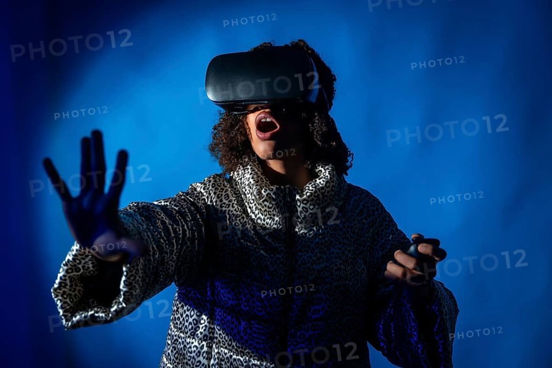 Excited young woman wearing vr headset and using controllers is experiencing metaverse on blue background in studio shot