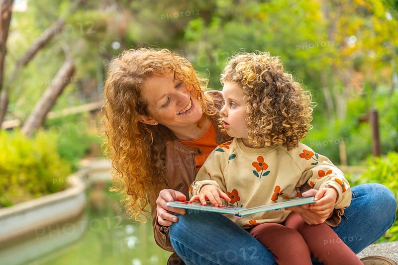 Cute girl and smiling caucasian mother reading a book sitting on a park together