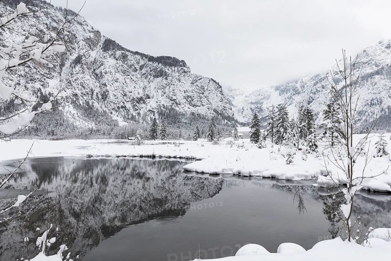 Snow covered Lake Almsee which is partly frozen and snow covered. Snow on the trees and mountains with gray overcast sky. Nearly a black and white scene