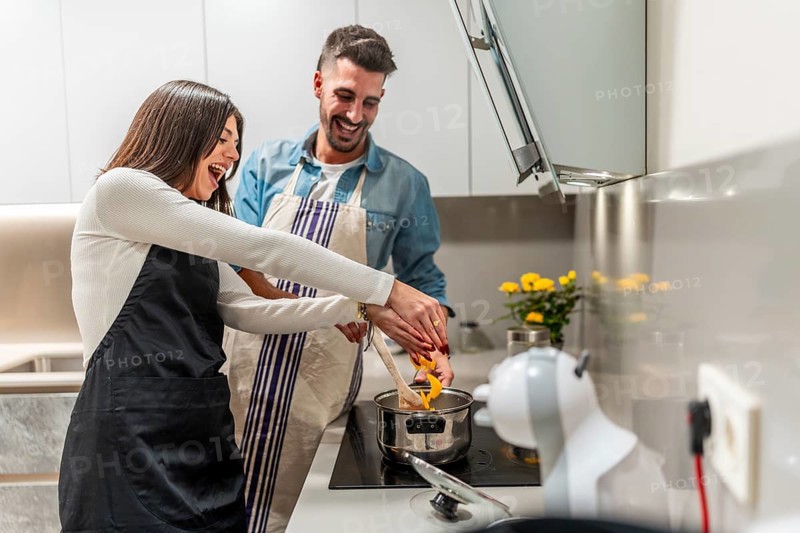 Happy couple wearing aprons preparing food in modern kitchen, stirring in pot and laughing