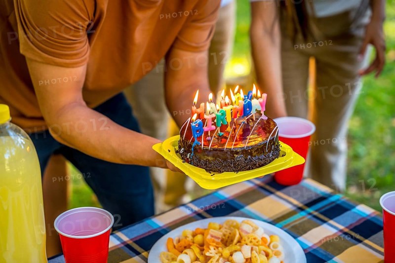 Close-up of the hands of a young man holding birthday cake with candles during picnic with friends