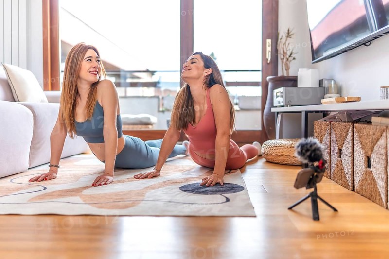 Two women recording an online yoga class from home, performing the cobra pose on a rug in the living room