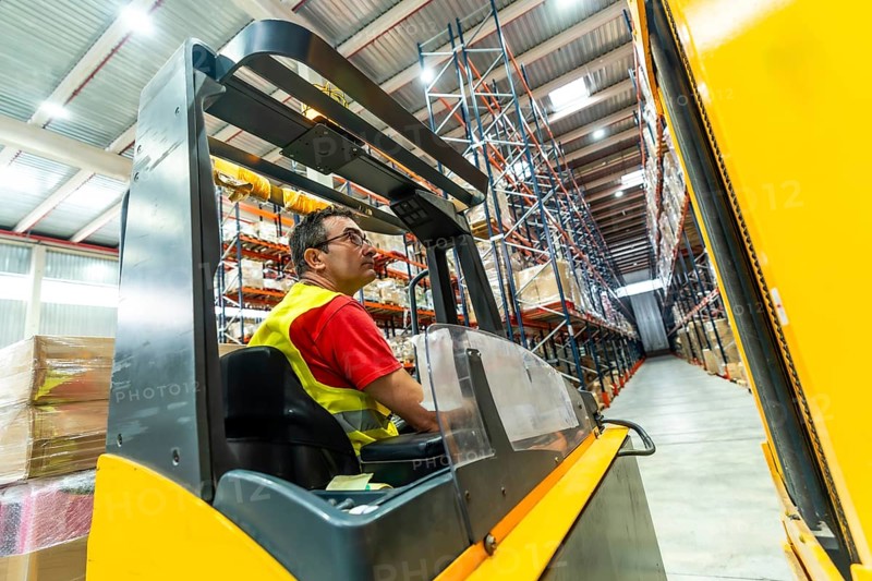 A worker in a yellow waistcoat is sitting driving a forklift in an industrial environment