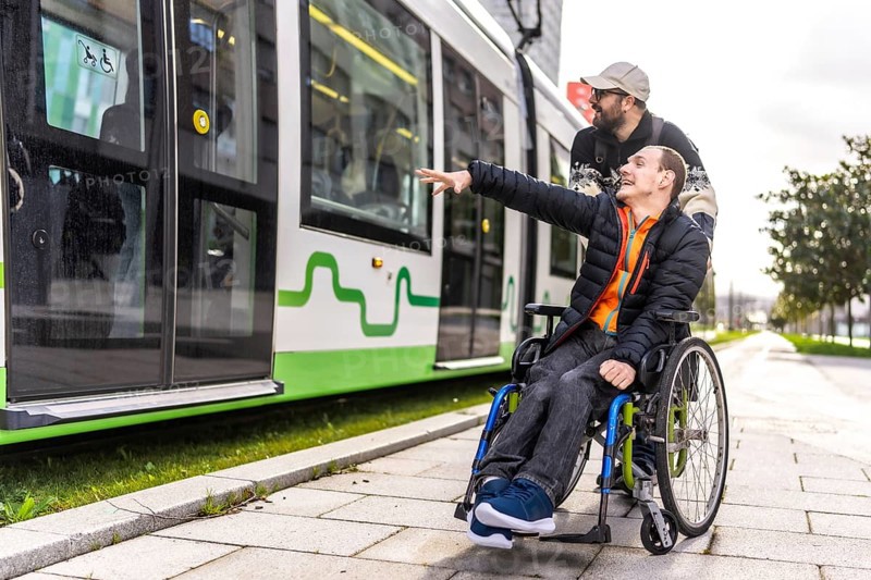 Young man in a wheelchair points at an approaching tram while his friend stands behind him, offering support and companionship