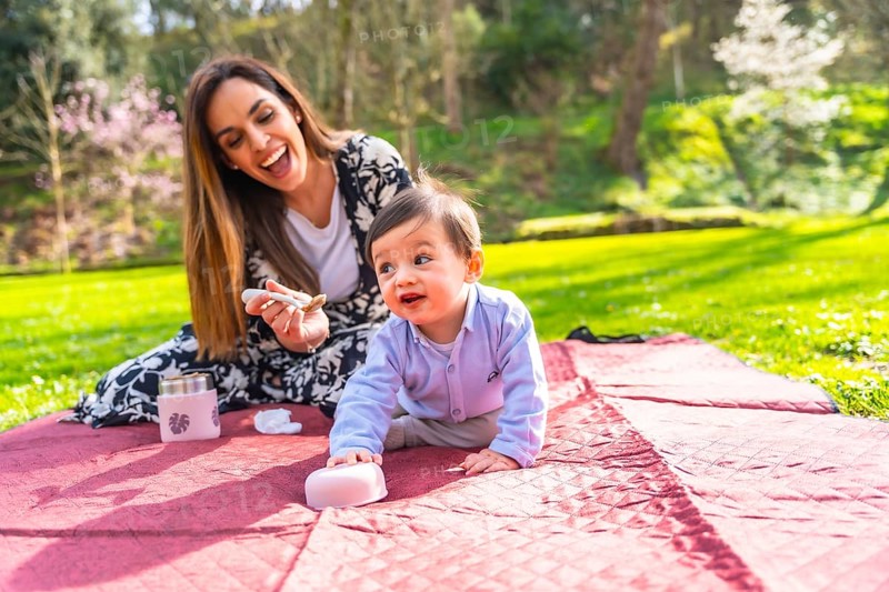 Mother giving food to her baby son during a picnic on a blanket in a park, surrounded by green grass and trees