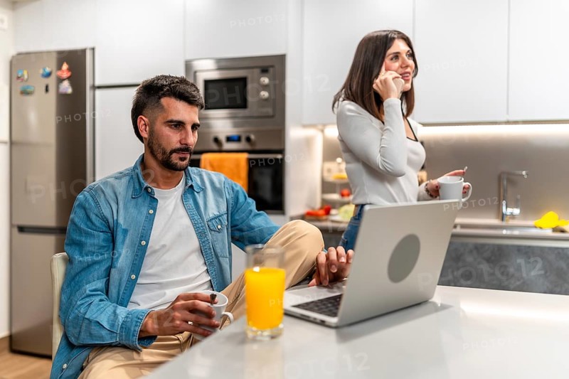 Young couple working from home in the kitchen, having breakfast and using laptop and smartphone