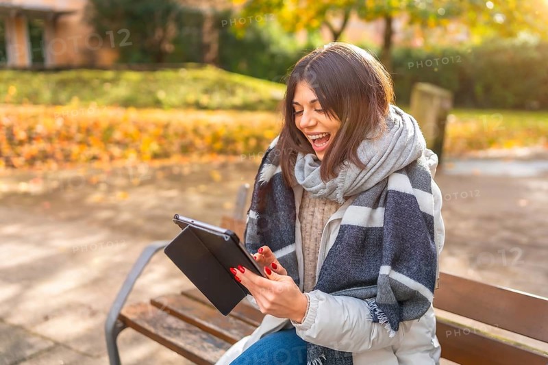 Rays of the sun illuminating a caucasian happy woman laughing while using digital tablet in a park in a cold day of autumn