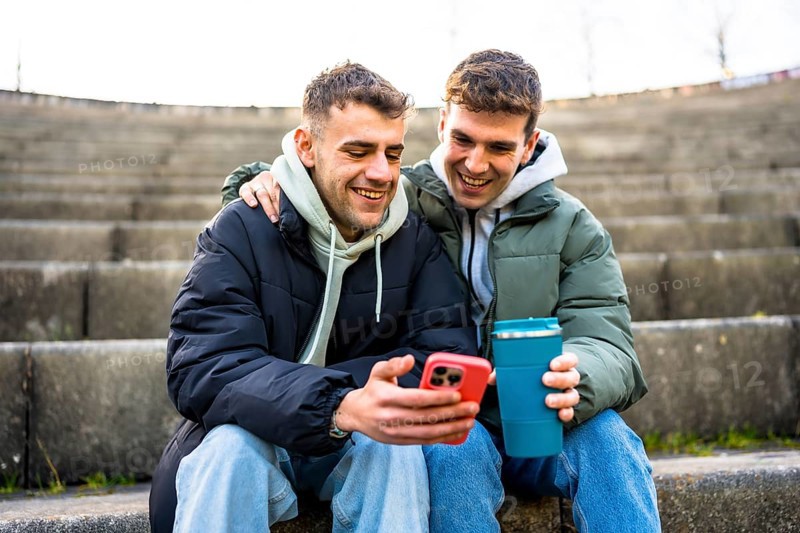 Two men are sitting on steps, embracing and looking at a smartphone while one holds a reusable coffee cup, enjoying a moment of togetherness