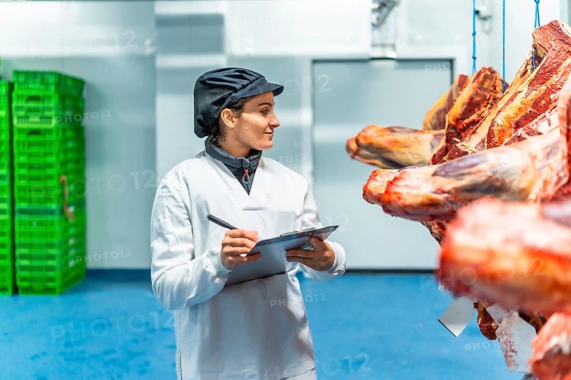 Woman in uniform and hat writing on clipboard checking goods in a meat processing factory