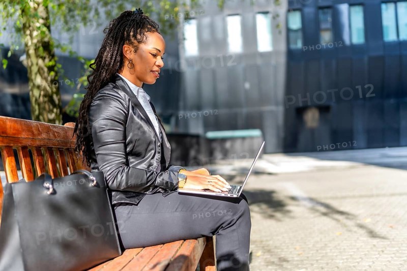Latin beauty and elegant businesswoman working on remote using laptop sitting on a bench sunbathing in the financial district