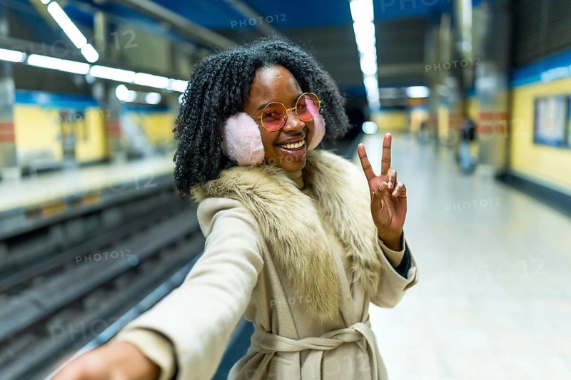 Smiling woman wearing earmuffs and pink glasses making selfie and victory sign while waiting subway train in station platform