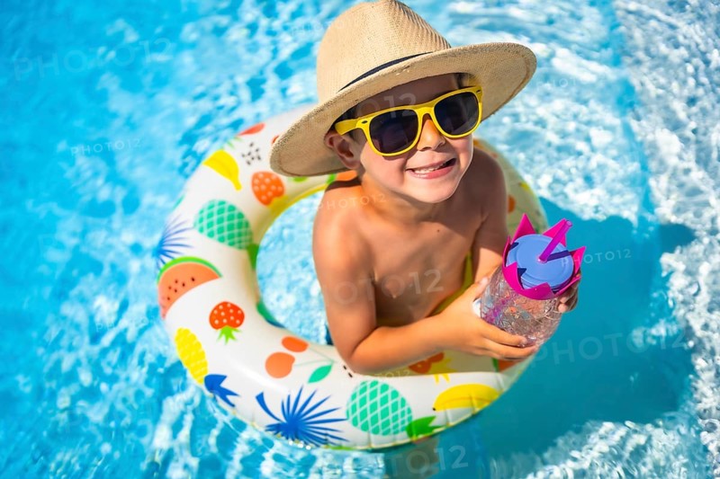 Top view close-up of a funny caucasian boy inside a pool drinking water into a rubber ring colorful float smiling at camera