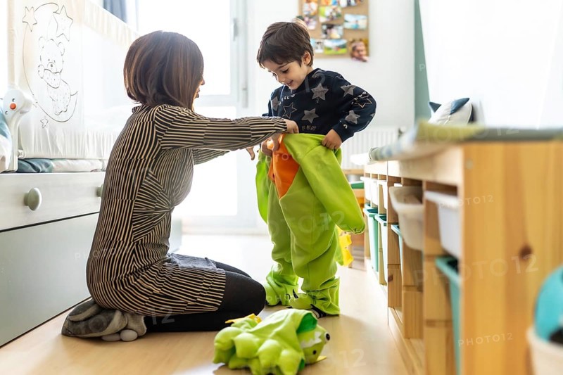 Mother assisting her son in putting on a colorful dinosaur costume, enjoying playful moments together in the cozy bedroom