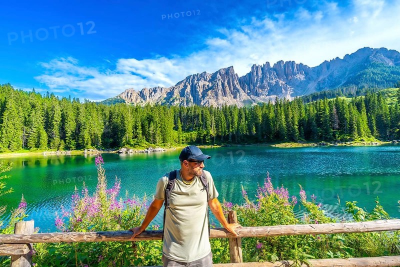 Hiker admiring the stunning turquoise waters of lake carezza with the latemar mountain range in the background, located in the italian dolomites