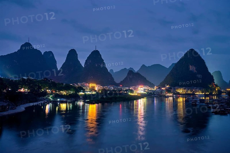 View of Yangshuo town illuminated in the evening with dramatic karst mountain landscape in background over Li river. Yangshuo, China, Asia