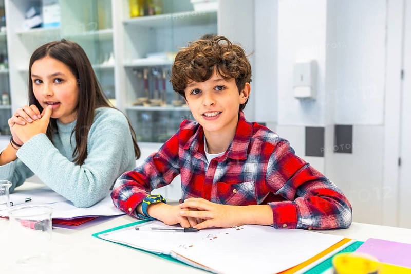 Smiling students sitting at desk in science classroom, learning and interacting during lesson, notebooks and educational materials visible