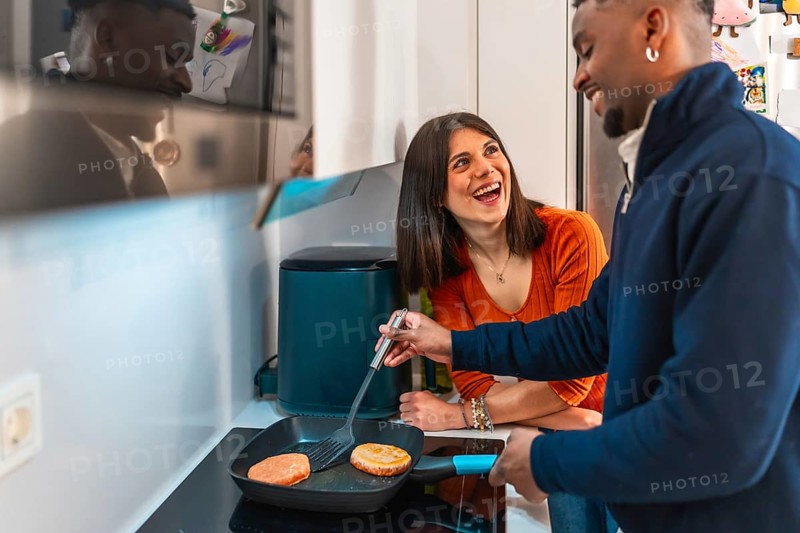 Cheerful multiethnic couple laughing and having fun while preparing burgers together on the stovetop in their modern kitchen