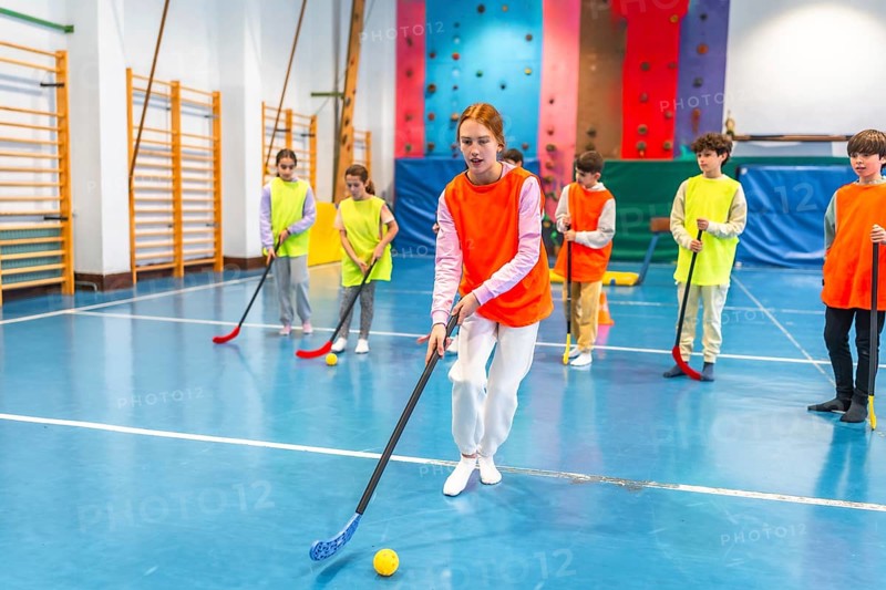 Group of elementary school students wearing orange and yellow vests playing floorball in a gym class, learning teamwork and sportsmanship