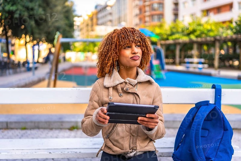 Pensive latin young woman in casual warm clothes using digital tablet sitting on bench in the city