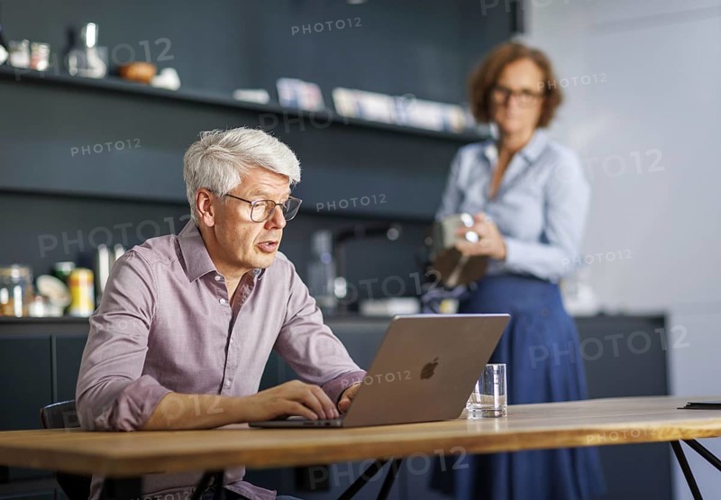 Symbolic photo on the subject of division of labour for couples in the household. A man sits in a kitchen at his laptop while a woman washes dishes in the background. Berlin, 13.08.2024
