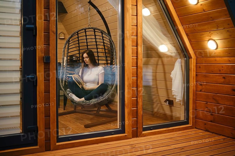 Cute woman sitting in a hanging swing chair and reading a book in the evening