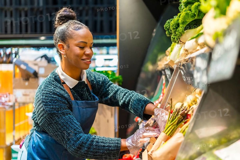 Smiling saleswoman wearing gloves arranging fresh organic asparagus on display in a supermarket