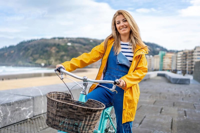 Smiling blonde woman wearing yellow raincoat and denim overalls riding bicycle with wicker basket along beach boardwalk