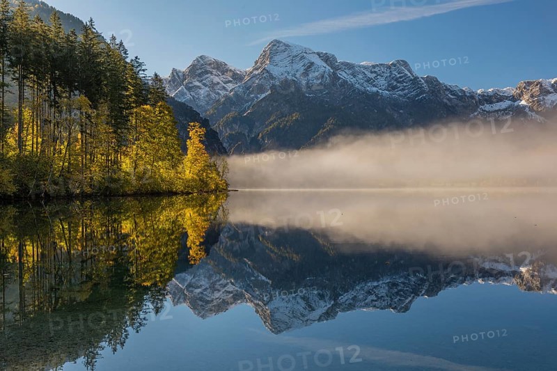 Lake Almsee in autumn with a forest and mountains reflected in the lake. A slowly dissipating fog moves over the lake. Blue sky