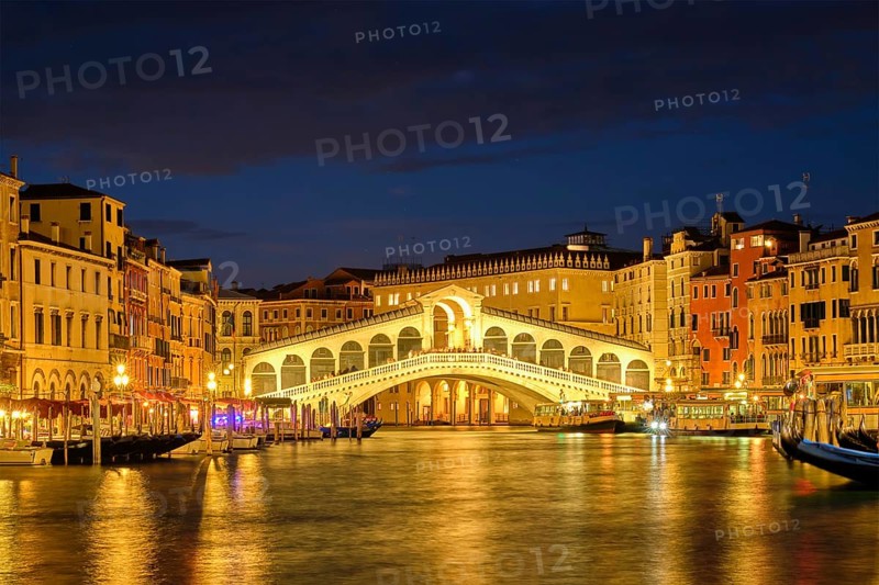 Famous Venetian tourist landmark Rialto bridge (Ponte di Rialto) over Grand Canal with vaporetto waterbus illuminated at night in Venice, Italy, Europe