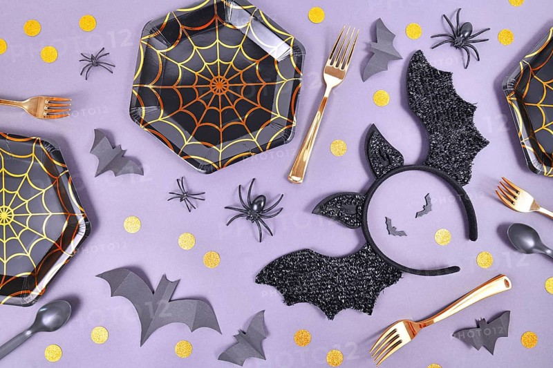 Halloween party flat lay with spider web plates, costume bat headband, spiders and confetti on violet background