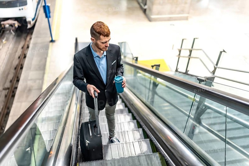 Young businessman going down an escalator in a station, carrying a suitcase and a coffee, going to work