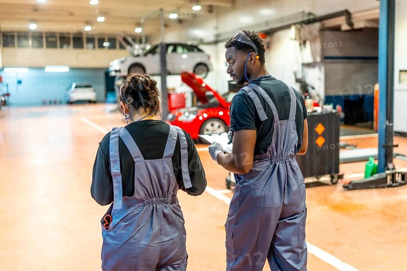 Two auto mechanics walking and reviewing notes on a clipboard in a professional garage, surrounded by vehicles on lifts