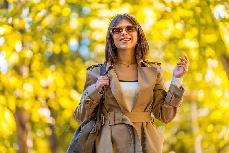 A woman wearing a tan coat and sunglasses is walking down a path. She is smiling and holding a handbag. Autumn or winter concept in the city