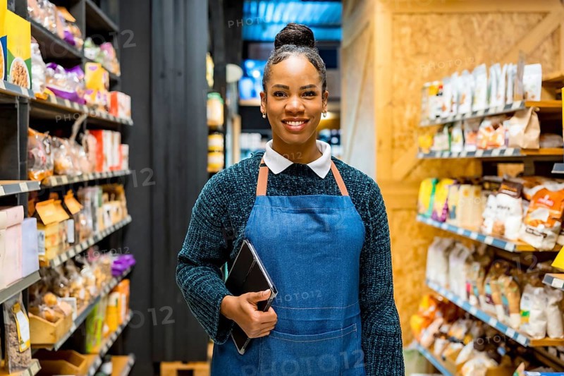 Confident saleswoman smiling and holding tablet, working in organic supermarket with shelves full of natural and healthy products