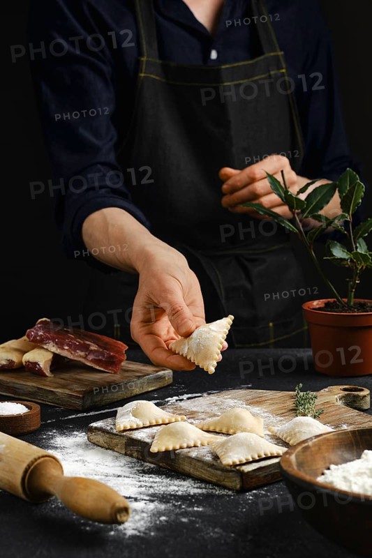 A person in an apron carefully shapes filled pasta in a warmly lit kitchen. Flour dust covers the countertop as various ingredients are arranged nearby, highlighting the process of handmade cooking.
