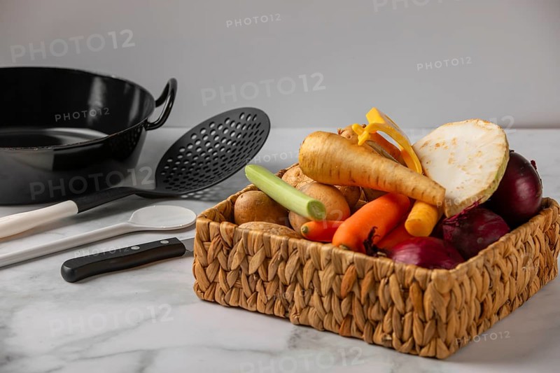 A woven basket full of vegetables such as soup vegetables, potatoes and onions next to kitchen utensils
